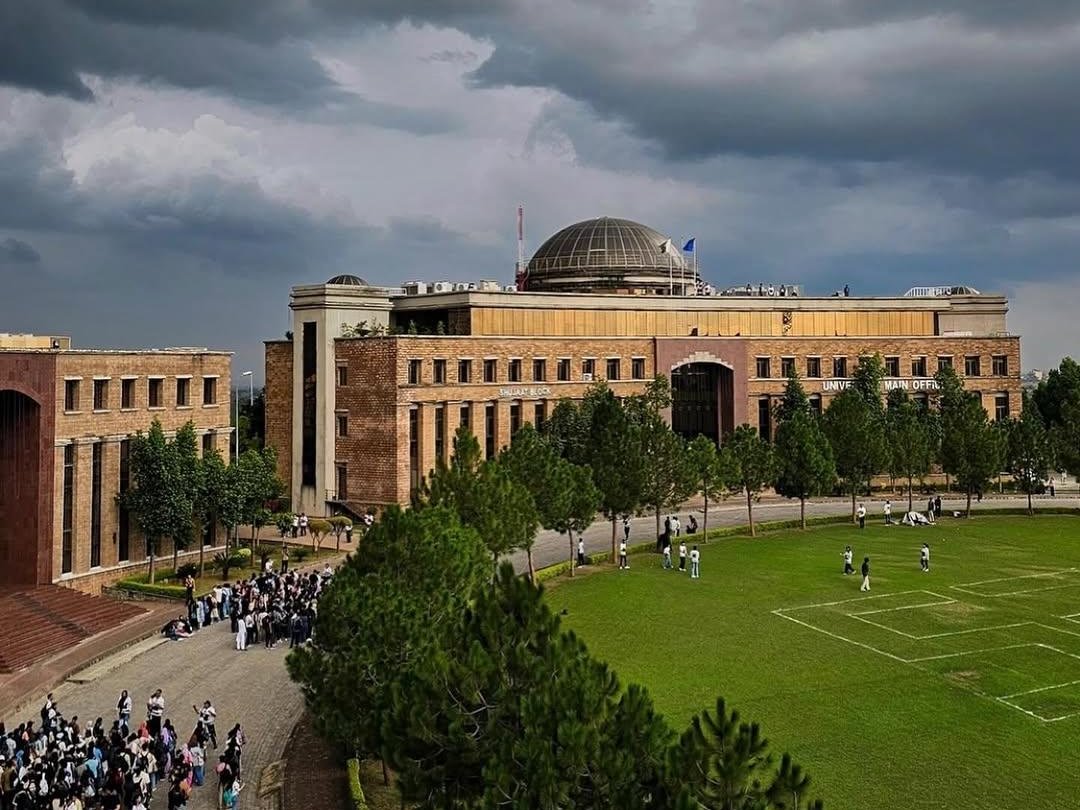 NUST Islamabad campus under storm clouds, with the main building, pine trees and playing field